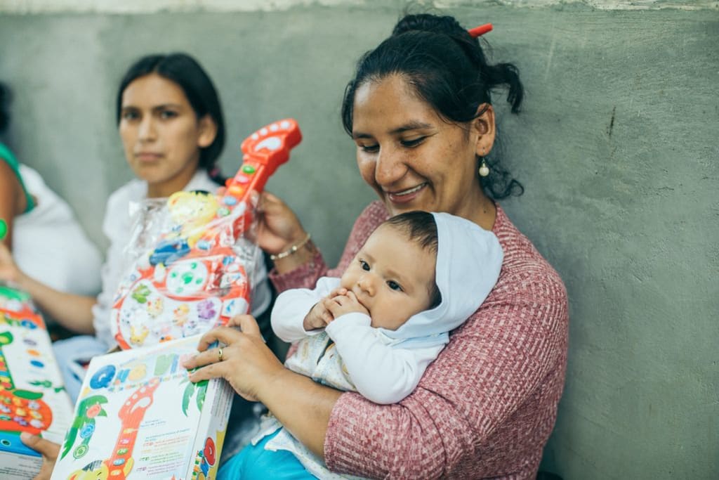 A mom holds her baby and a new toy guitar. She is wearing a pink sweater and the baby is wearing a blue sweater with a hood.
