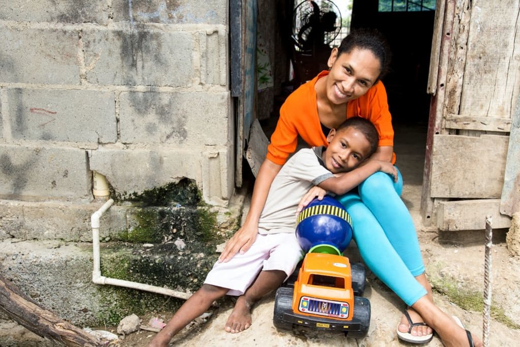 A mom sits beside her son who is holding her knees. He has a toy truck in front of him.