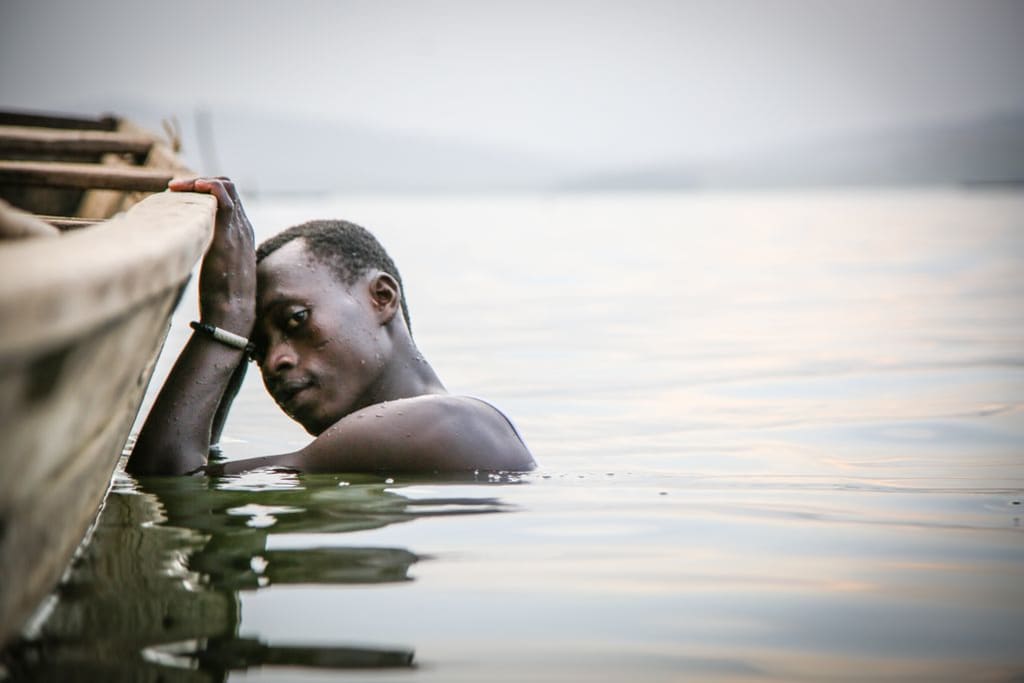 Boy in submerged in the water, his hands are holding onto the edge of the boat, as he looks at the camera with a defeated look in his eyes.