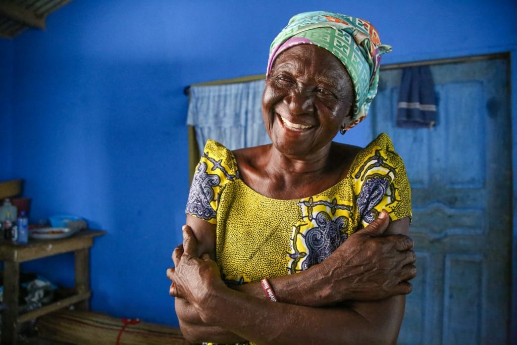 A woman in a yellow dress smiles in a dark blue room, with her arms crossed.
