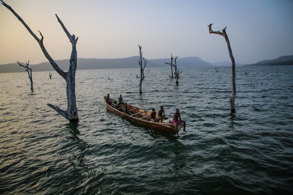 A group of boys sit in a wooden boat on choppy waters, surrounded in dead trees sprouting through the water.