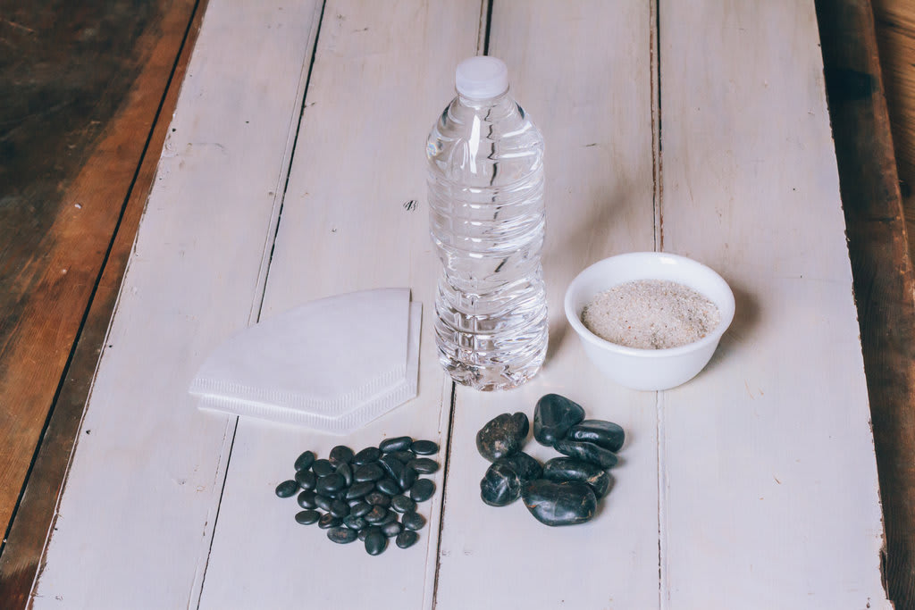 A water bottke, two piles of rocks, a bowl of sand and two coffee filters sit on a wooden table.