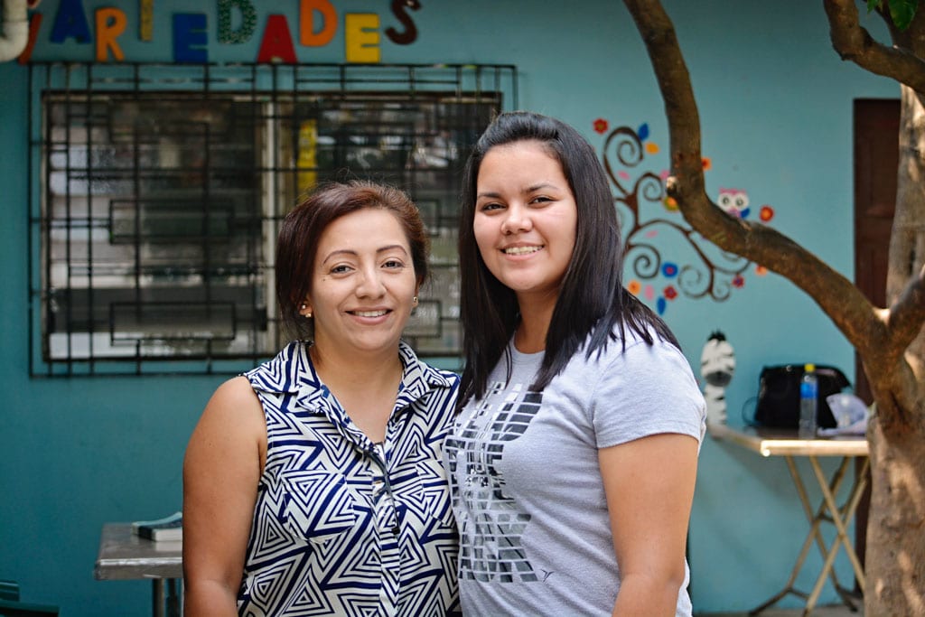 A young woman stands next to her former tutor. They stand in a colourful classroom.