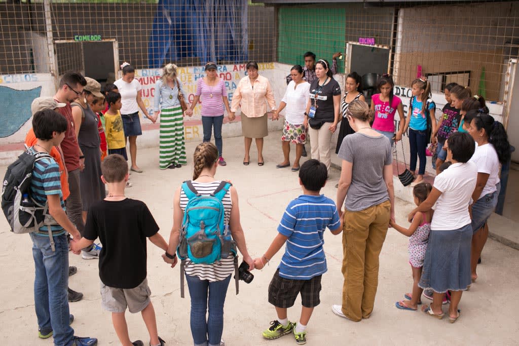 Visiting sponsors, visitors, adult men and women, parent, mothers and fathers, with their children, sons and daughters, families, stand together in a large circle holding hands and praying, prayers, with each other during a prayer time at the center.