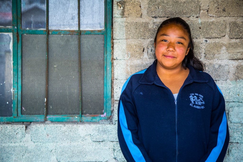 A teen girl stands against a wall beside a window smiling. She is wearing a blue zip-up sweater.