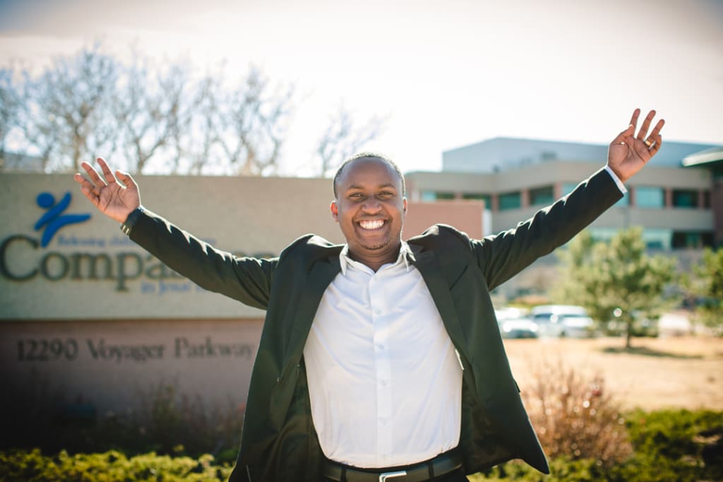 Jean Claude is pictured in front of a Compassion sign. He is smiling with his arms raised and is wearing a suit.