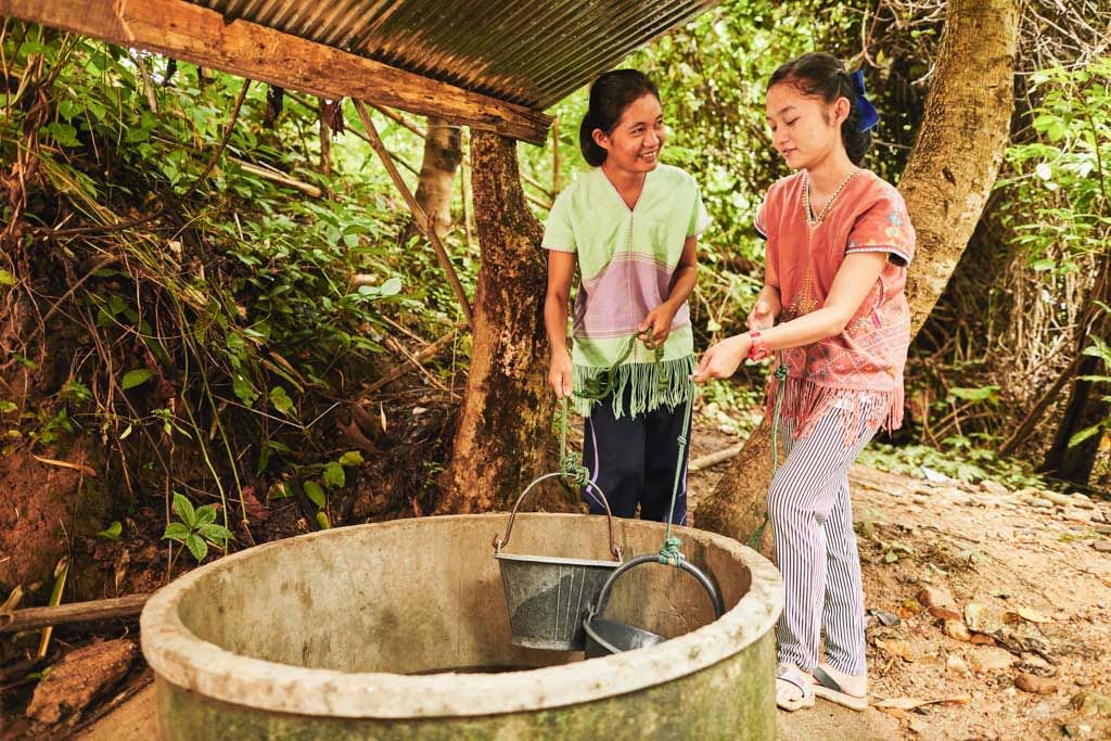 One woman and one girl are pictured fetching water with their pails from a well. They are under a shelter with a tin roof.