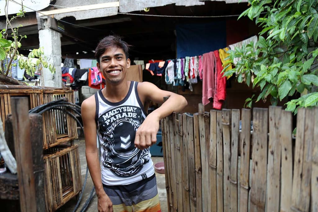 A young man leans on a fence outside his house and smiles.