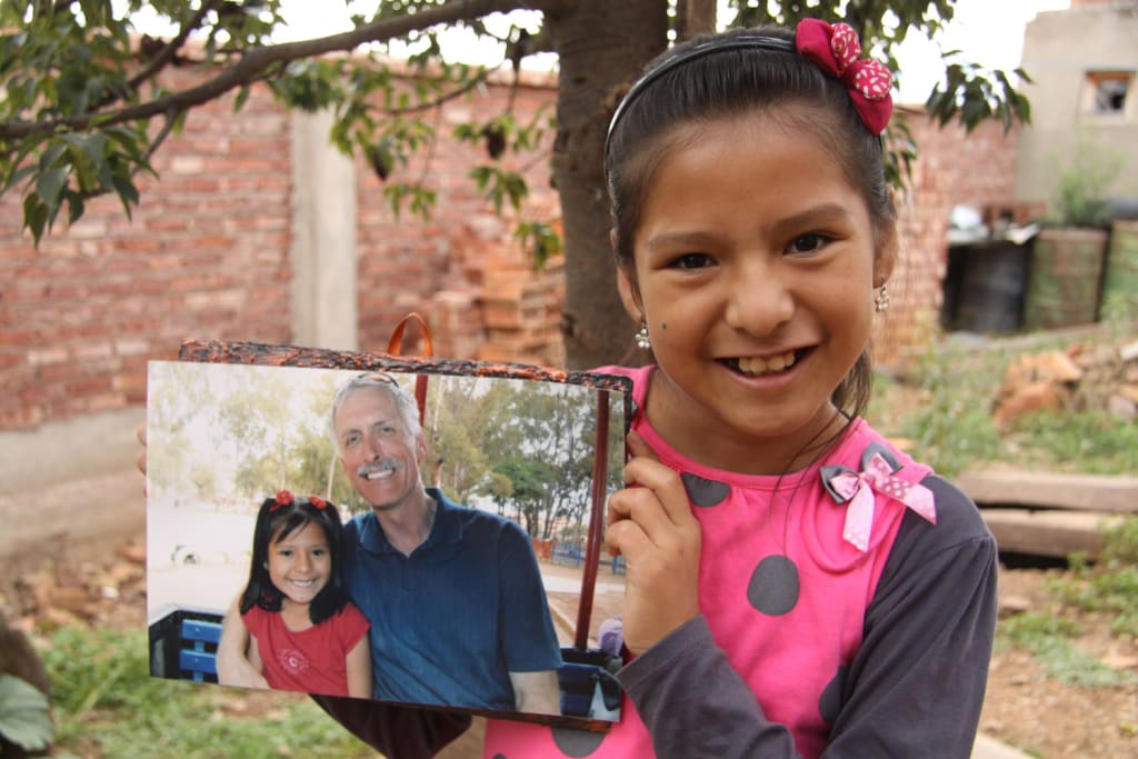 Girl wears a pink bow in her hair and a matching pink jumper. She is holding up a picture of her and her sponsor.