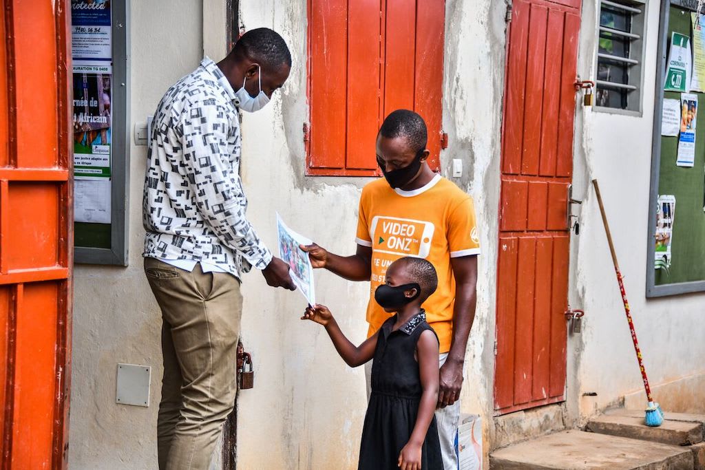 Fortune is wearing a black dress and a face mask. She is standing at the entrance to the centre with her father, wearing an orange shirt and a black face mask, and Tonton, the centre's social worker, who is handing Fortune a letter from her sponsor.