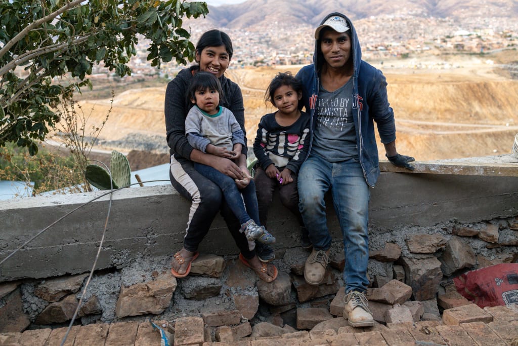 Ruliana sits with her husband and two daughters on a small wall near a tree. They all smile.