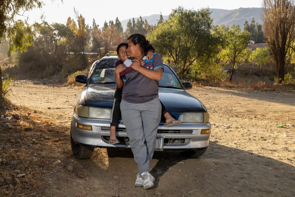 A woman is standing in front of her taxi with her son, who is sitting on the hood of the car and hugging his mom.
