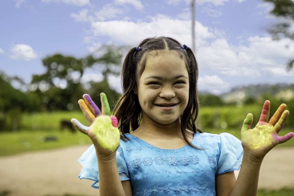 Girl wears a blue dress and smiles holding up her hands that are covered in paint.