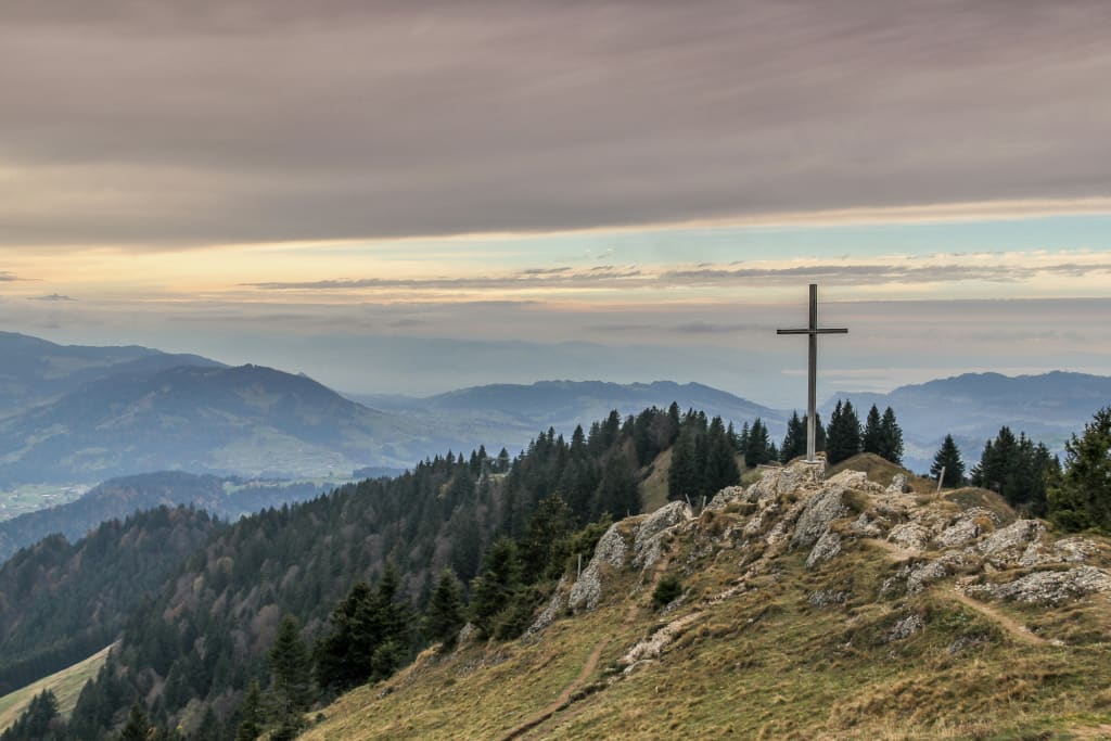 A cross stands on a hill overlooking forested hills.