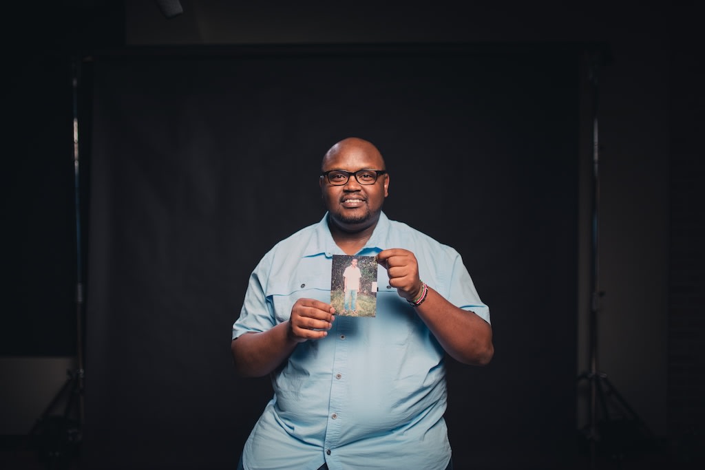 Owen, an alumni from Kenya, is wearing a light blue shirt. He is holding up a picture of himself as a child