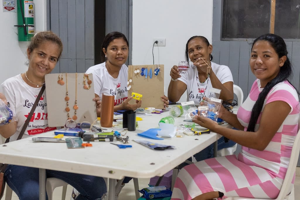 A group of women sit at a table and show their recycled jewelry to the camera. They are all smiling.