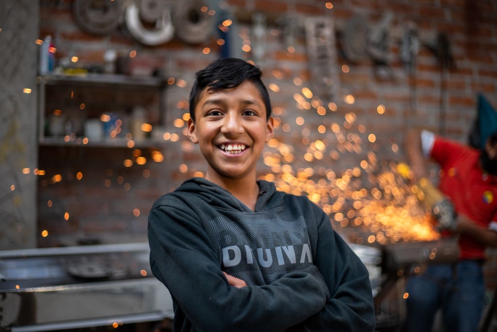 In Ecuador, 13-year-old Cristofer is wearing a dark gray sweatshirt and tan pants. He is standing inside an industrial machine shop with his arms crossed in front of him. There are sparks in the background.