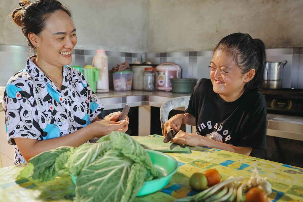 Karunia is smiling while cutting vegetables with her mom, Angel. There is lettuce in front of them in a bowl.