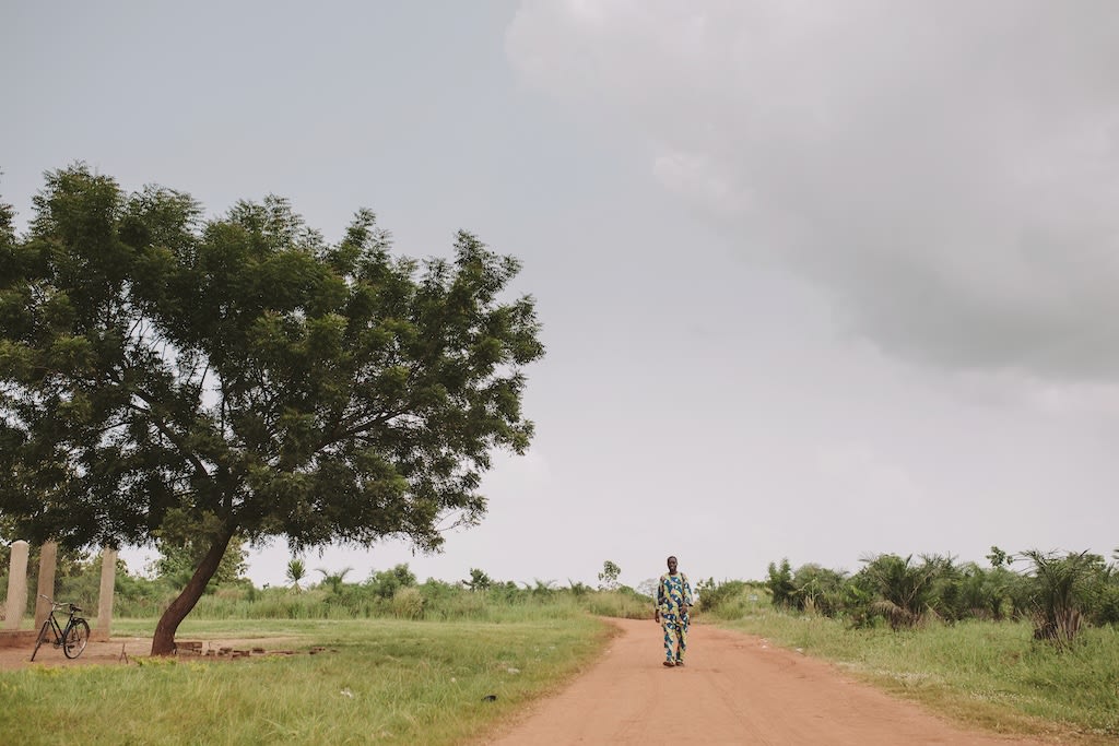 A man walks down a dirt road with a large tree on the side.