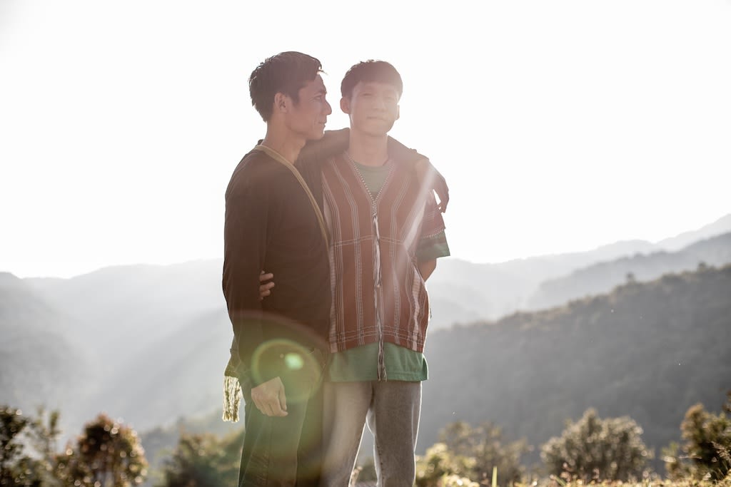 Two men standing on a mountain with the sun shining down on them. One has his arm over the other's shoulder.
