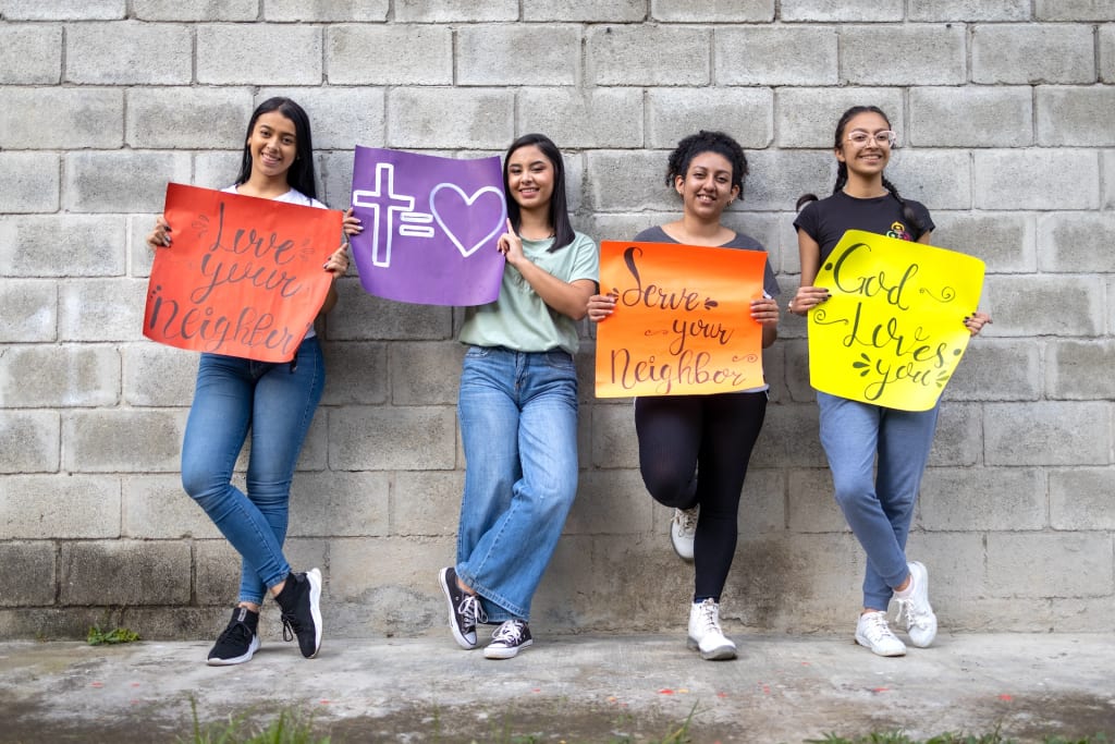 Four girls stand against a wall and hold up signs that say "Love your neighbour" and "God loves you".