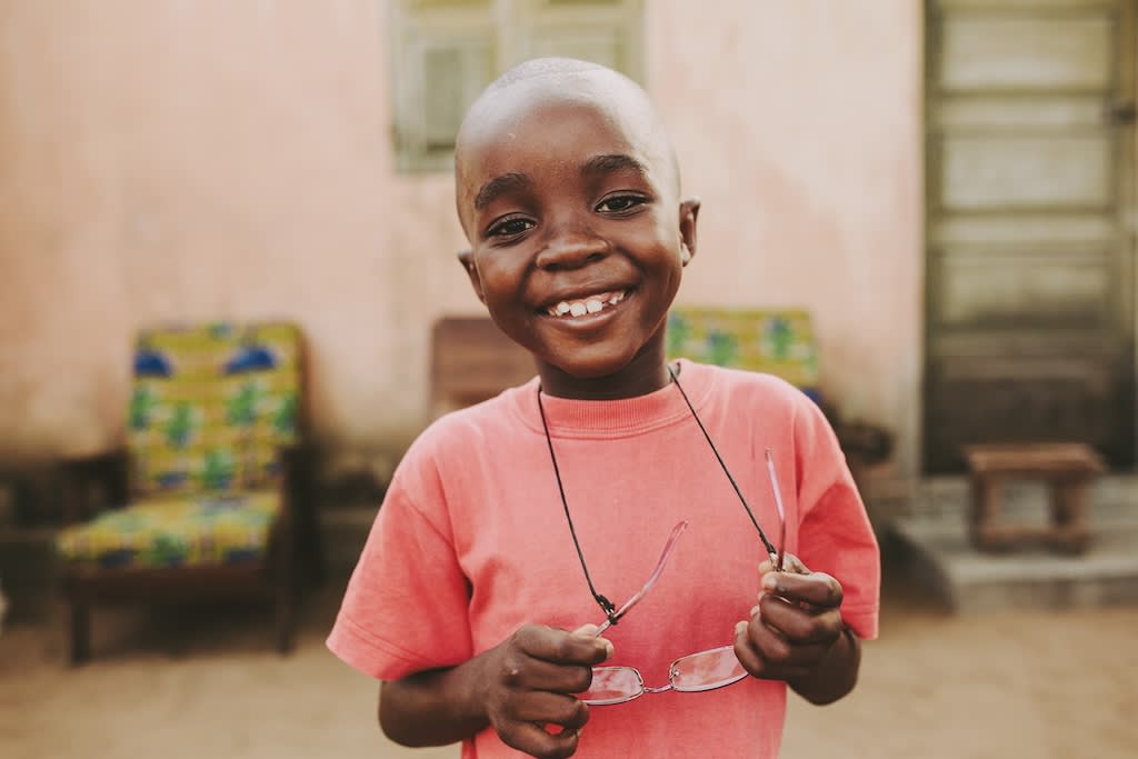 A young boy with a pink shirt smiles at the camera with glasses around his neck.