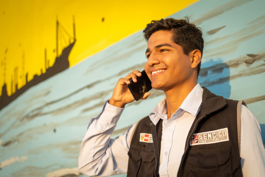 A young man in a vest holds a cellphone to his ear and smiles. He stands in front of a yellow and blue wall.
