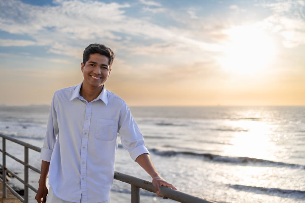 A young man in a blue shirt stands in front of the ocean during a sunset.