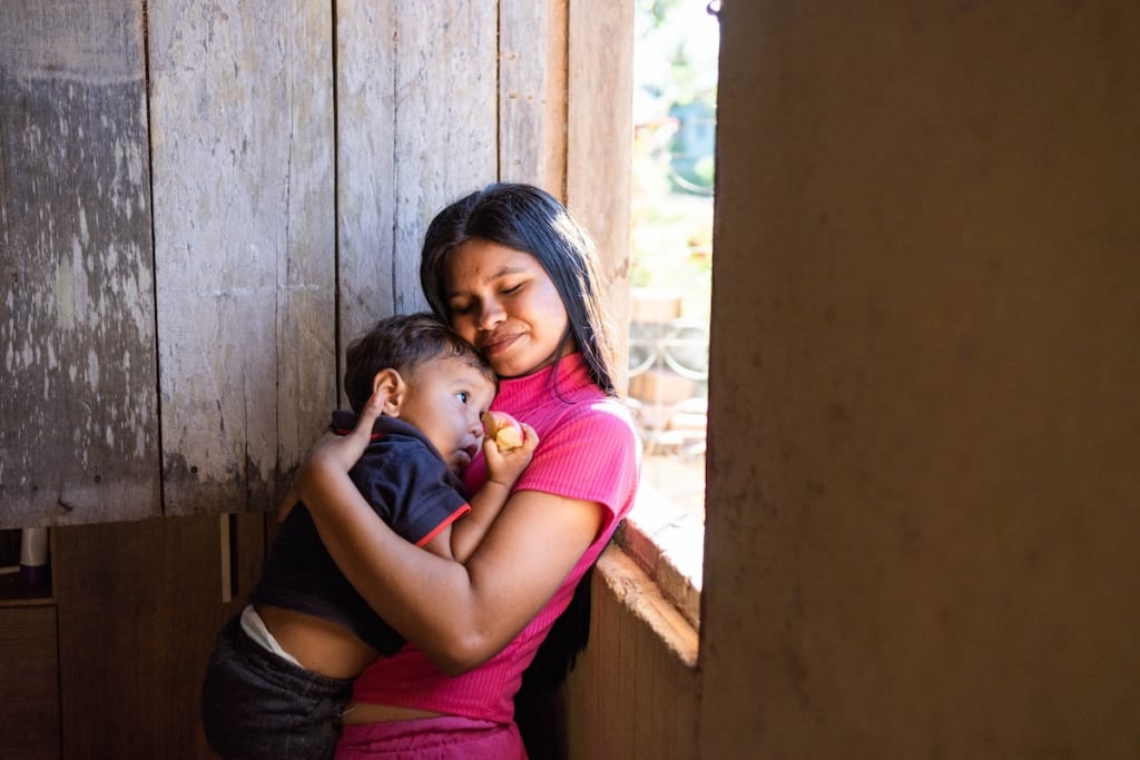 Gabriella is holding her son, Ithallo, while standing at the front window of her house.