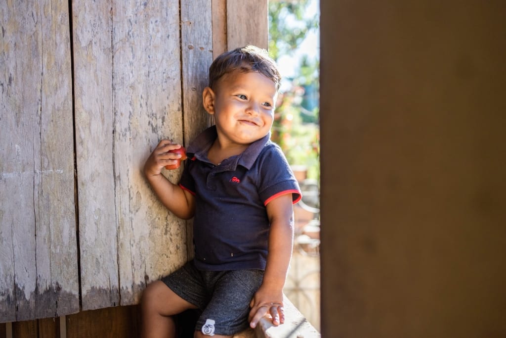 Gabriella's son, Ithallo, is smiling as he sits in the window eating an apple.