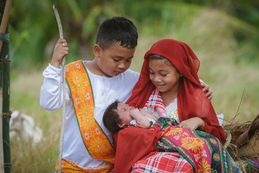 A girl dressed as Mary holds a baby, playing Jesus, in her lap. A boy dressed as Joseph has her arm around Mary and looks down at the baby.