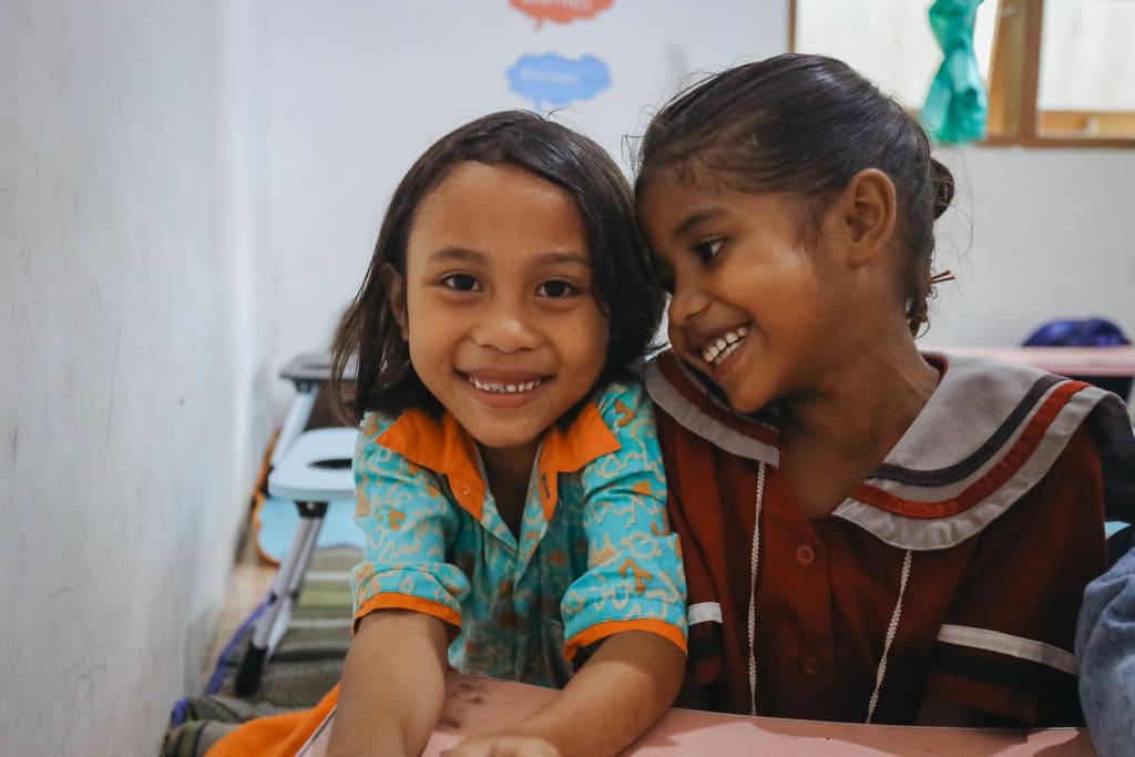 Two young girls in colourful dresses sit smiling at a desk.