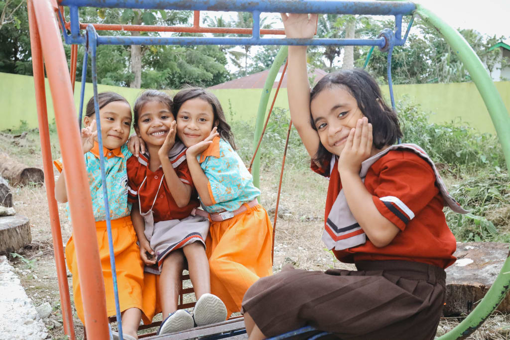 Four girls in colourful dresses sit on playground equipment and smile.