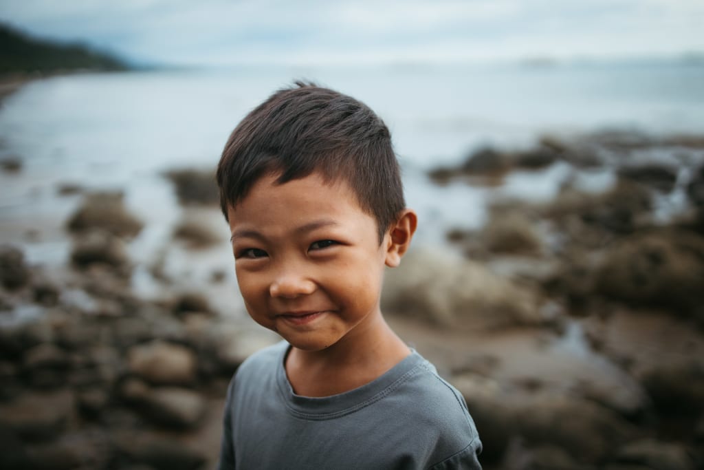A young boy stands in front of the ocean and smiles.