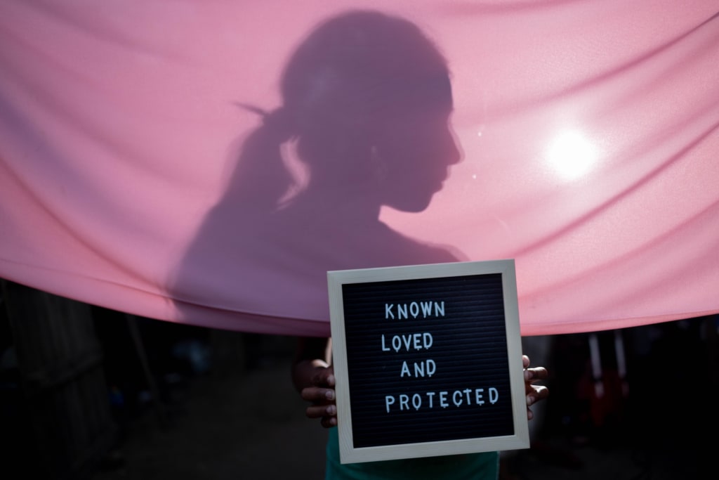 A person hidden by a pink curtain holds up a board that says "Known, Loved and Protected".