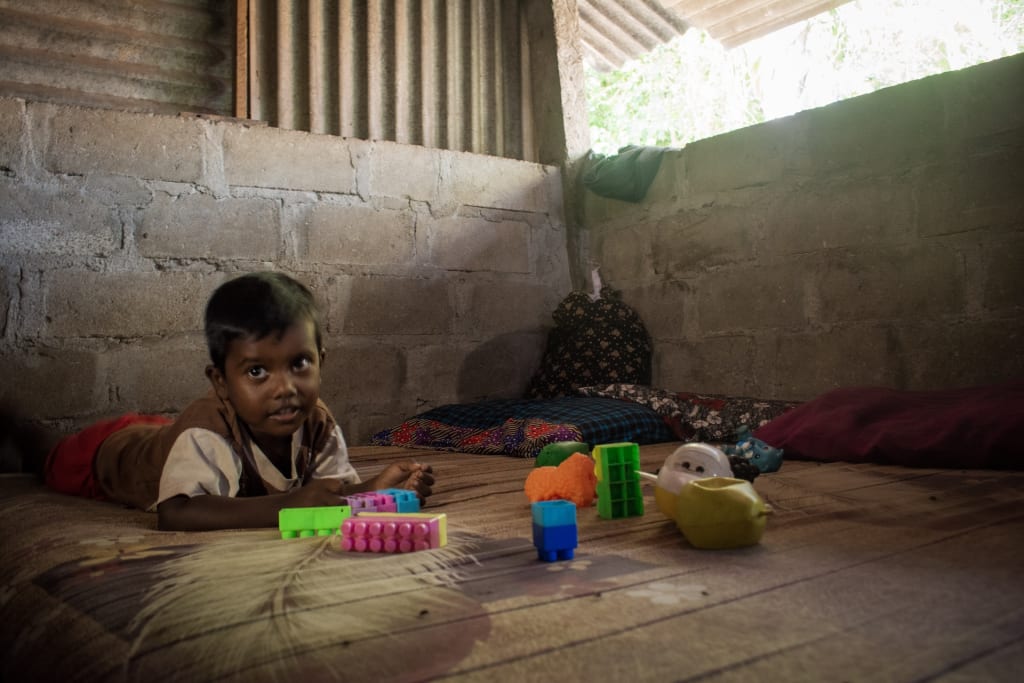 Rekonshan is wearing a brown shirt with tan sleeves and red shorts. He is laying down on the floor in his home and is playing with toys.