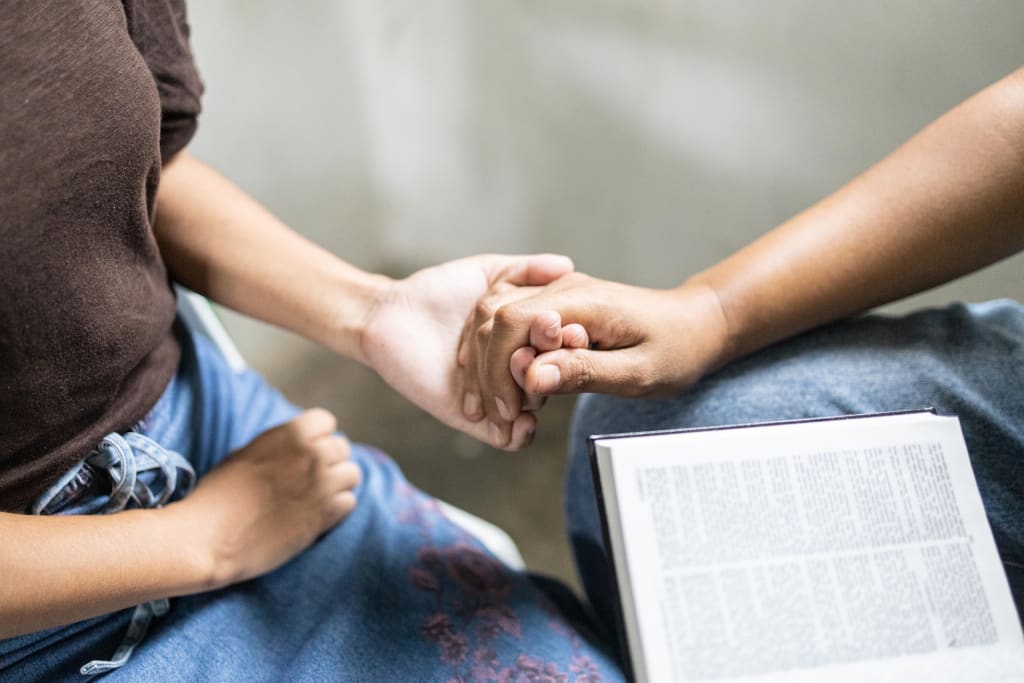 Closeup of Janna, in a brown shirt and blue denim skirt, sitting in a classroom at the center holding hands with Hazel, her child development worker. Hazel, in blue jeans, is a Compassion alumna. Hazel has a Bible open in front of her.