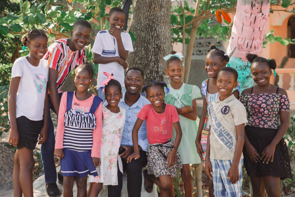 A man sits in front of a tree with a group of children in colourful clothing. They are smiling.