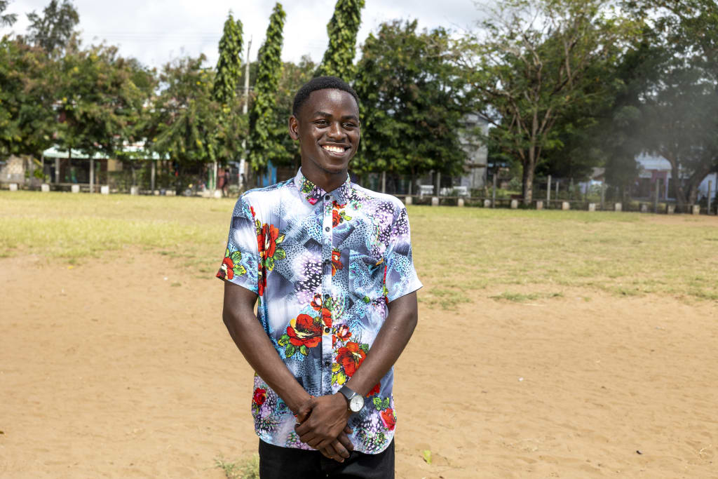 A teen boy stands in a patterned shirt with his hands clasped in front of him and smiles.