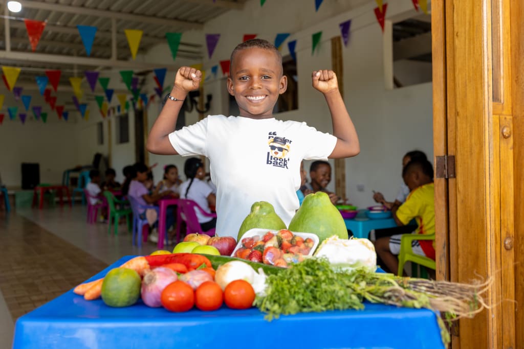 A boy holds his arms up and smiles. There is a table of fruit and vegetables in front of him.
