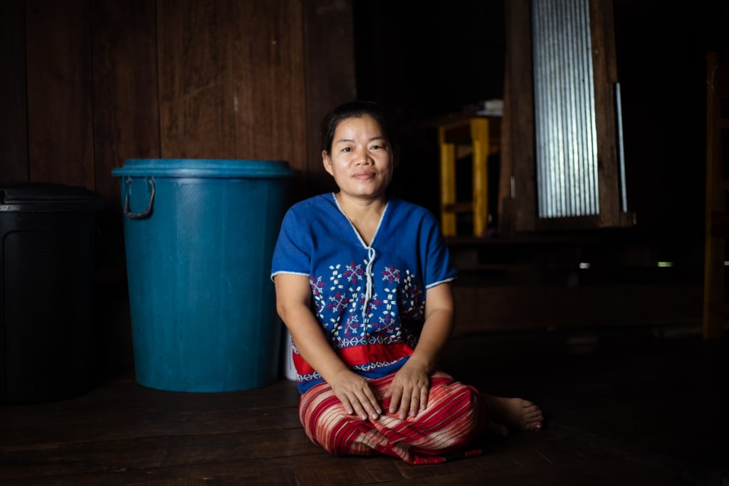 A woman kneels in a dark room. She wears blue and red. There is a blue bucket behind her.