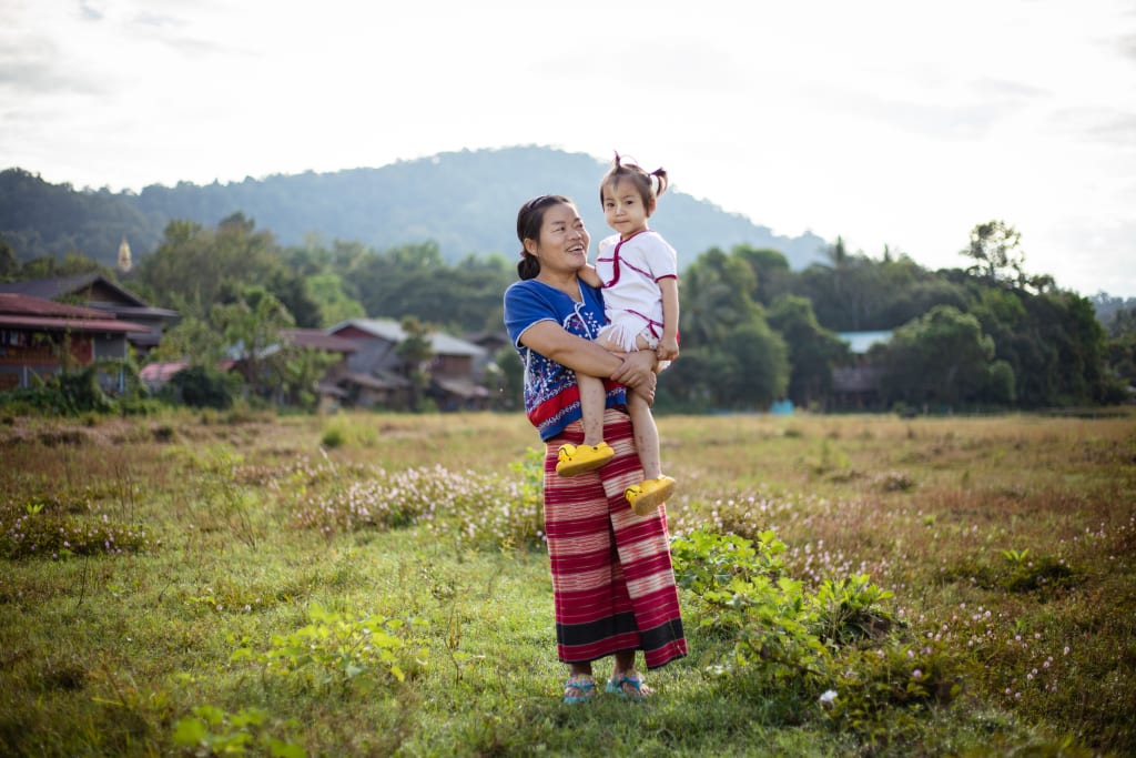A woman in blue and red holds a little girl in white. They are standing in a field with a large hill in the background.