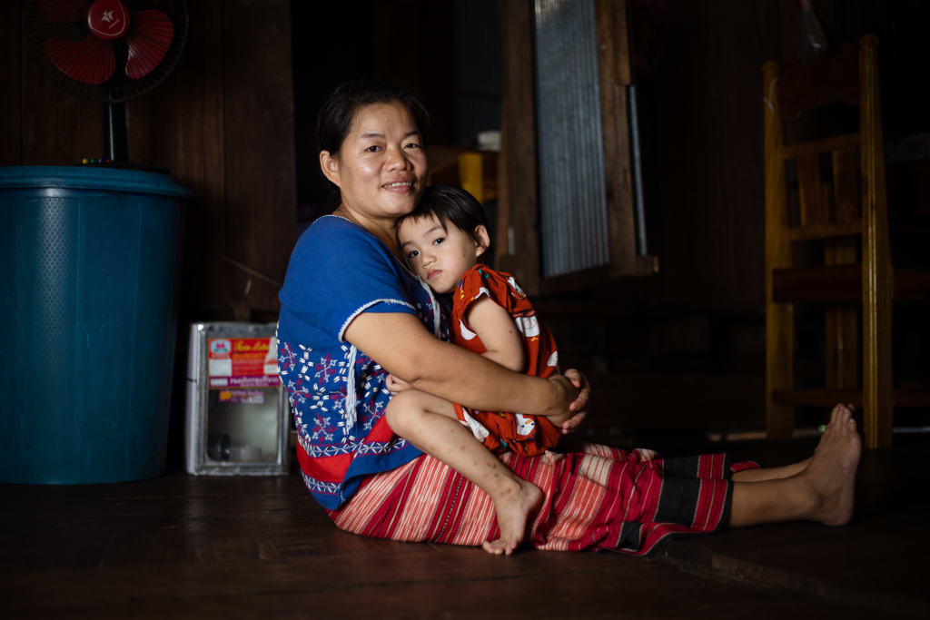A woman in a blue and red dress sits on the ground with her legs out. Her daughter wears a red dress and leans against her chest.