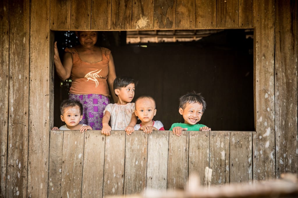 Four children are looking out the window of a wood building. A woman in an orange shirt and purple skirt stands behind them.