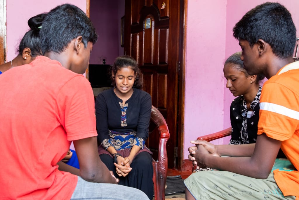 Ajey is wearing an orange shirt and Arun is wearing a red shirt. They are praying with the rest of the leadership team of the youth club that was formed through the local Compassion program.