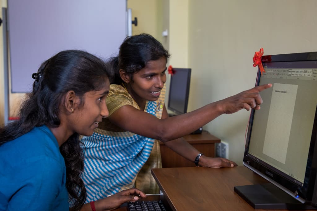 Ramyapriya is wearing a blue and white dress. She is sitting at a desk inside the new computer lab and is working on one of the computers while her tutor is teaching her.