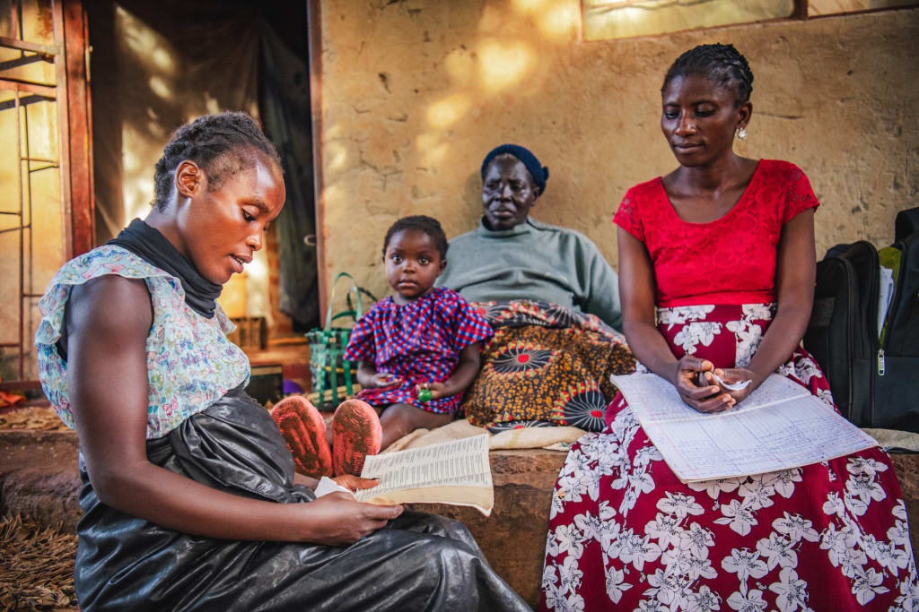 Gracious is reading the Bible with two other woman and her daughter Mayamiko in front of a brown building. Mayamiko, wearing a red and blue printed dress, is sitting next to a woman in a gray top. Next to her is a woman in a red top and red printed skirt holding a white paper in her lap.