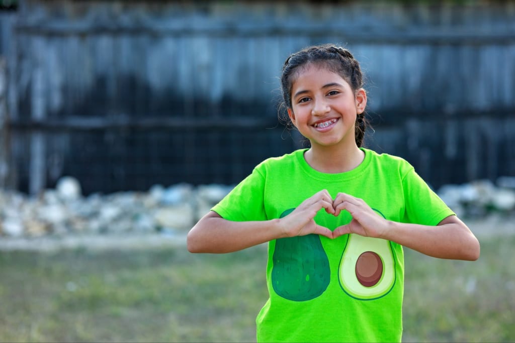 A young child smiles while making a heart with her hands.
