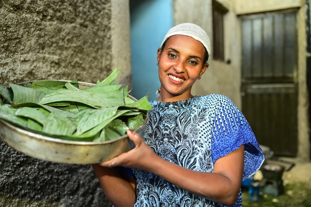 Tsehaynesh holds wheat dough in a baking tray that is covered with banana leaves. She is wearing a dress with blue print and a white scarf in her hair. She smiles at the camera.