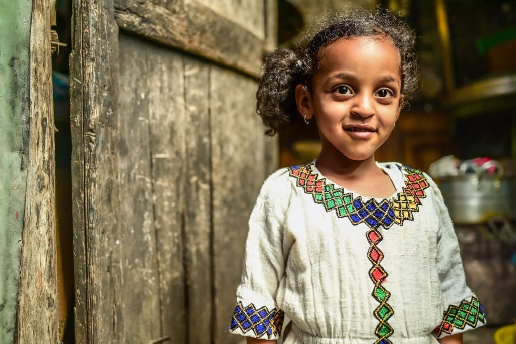 Elbetel stands in front of a wooden door in her home. She is wearing a white dress with a colorful geometric pattern.